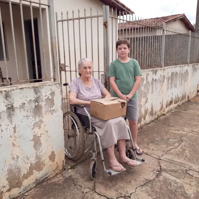 Neusa em frente ao portão de casa, com a caixa de doces no colo, ao lado do neto Mateus